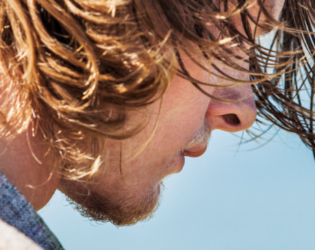 Man with long hair, standing in front of a body of water, with wind blowing in his hair.