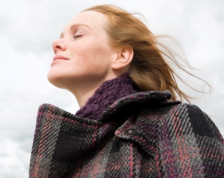 Woman with eyes closed, standing outside with a coat on, with wind blowing in her hair.  