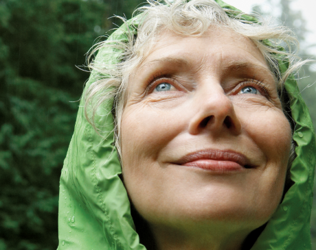 Woman wearing a green rain jacket, standing in a forest and looking up at the sky.